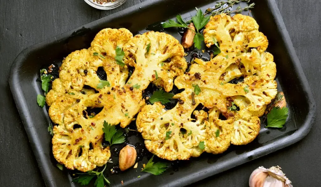 Baked cauliflower steaks with herbs and spices on baking sheet over black stone background.