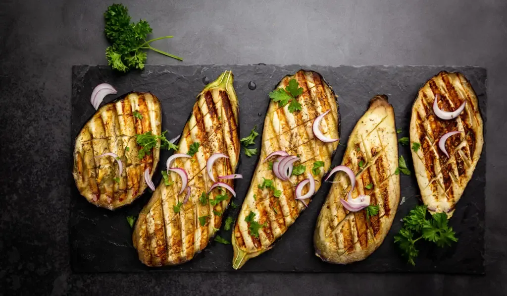 Roasted eggplant slices garnished with fresh herbs and onion on black background